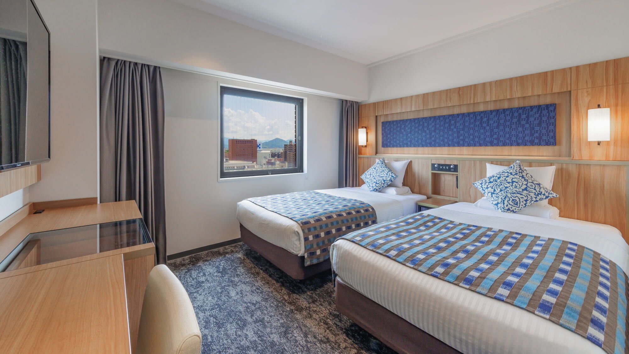 A hotel room with two beds, a desk, a TV, and a window with a city view.