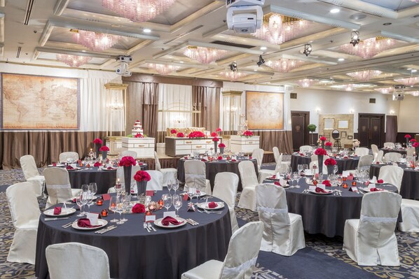 Un salón de banquetes con mesas redondas dispuestas para comer, sillas blancas y centros de mesa florales rojos.