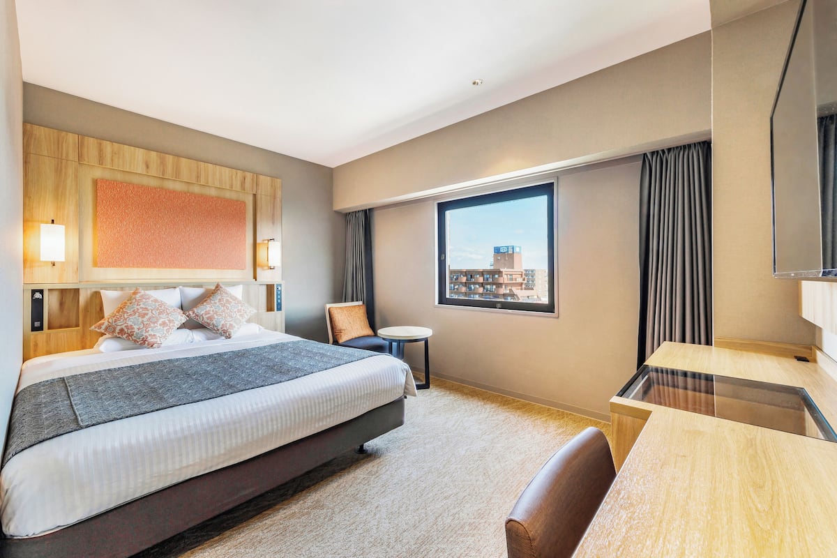 Quarto de hotel com cama grande, escrivaninha, cadeira, mesa redonda pequena e uma janela ampla com vista para um edifício.