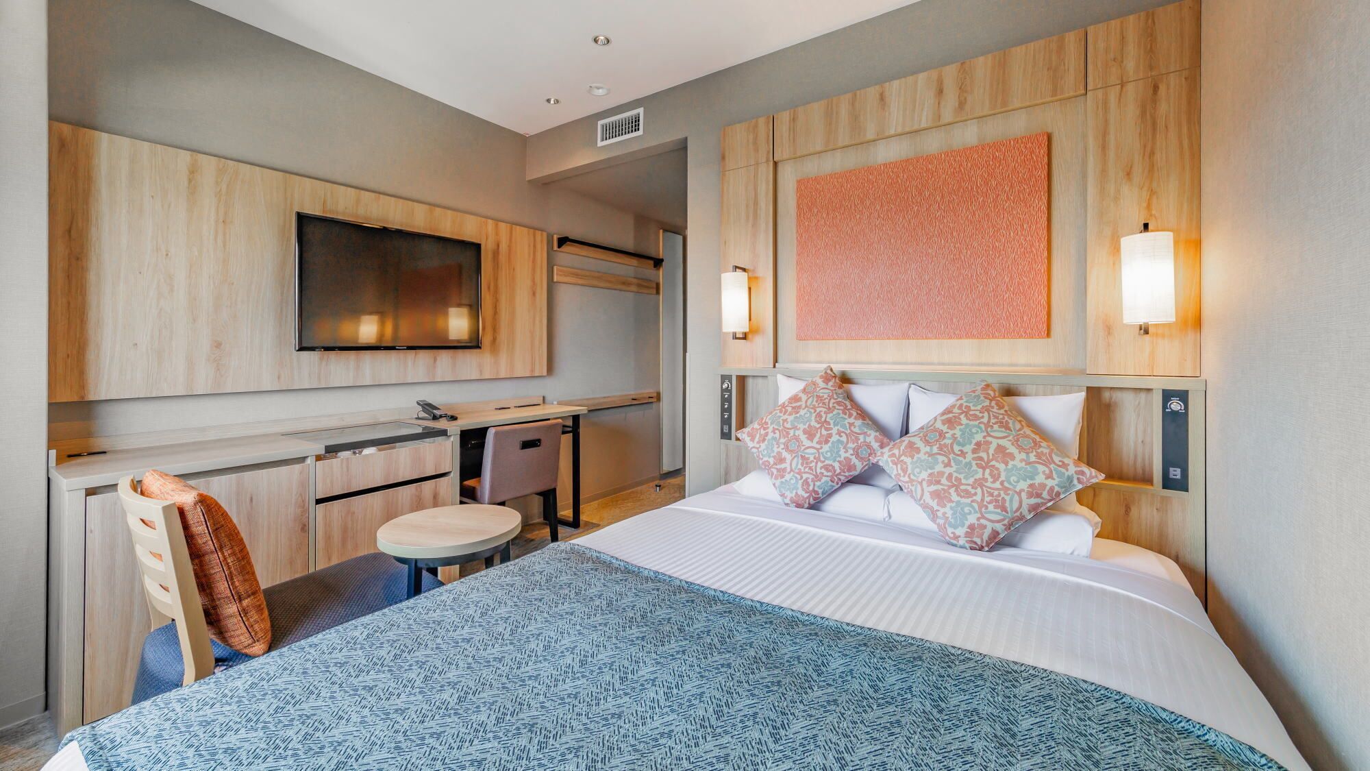 A hotel room with a bed, a desk, a chair, a TV, and a wall-mounted light.
