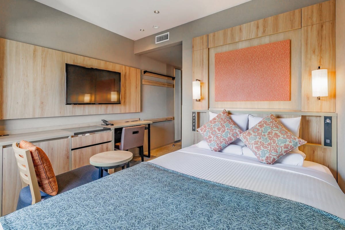 Quarto de hotel com cama, escrivaninha, cadeira, televisão e luminária na parede.