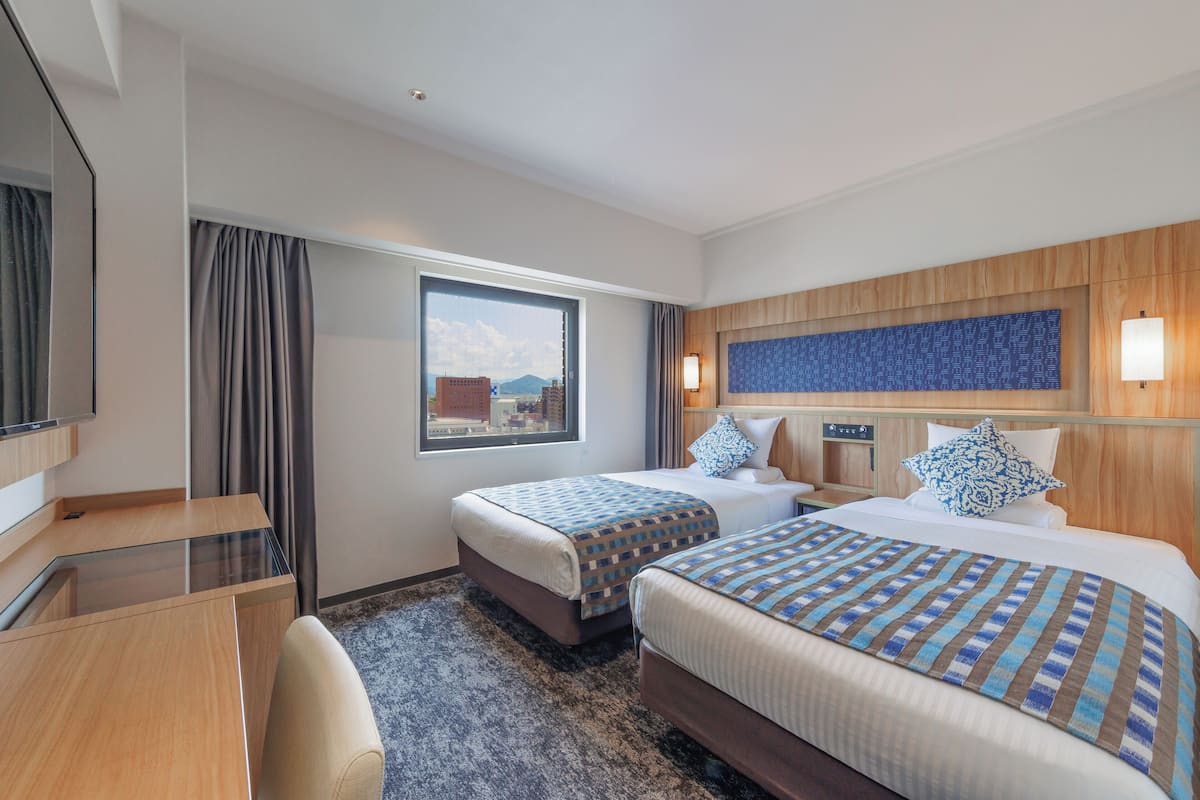 Quarto de hotel com duas camas, uma escrivaninha, uma cadeira, janela com cortinas e vista para a cidade.