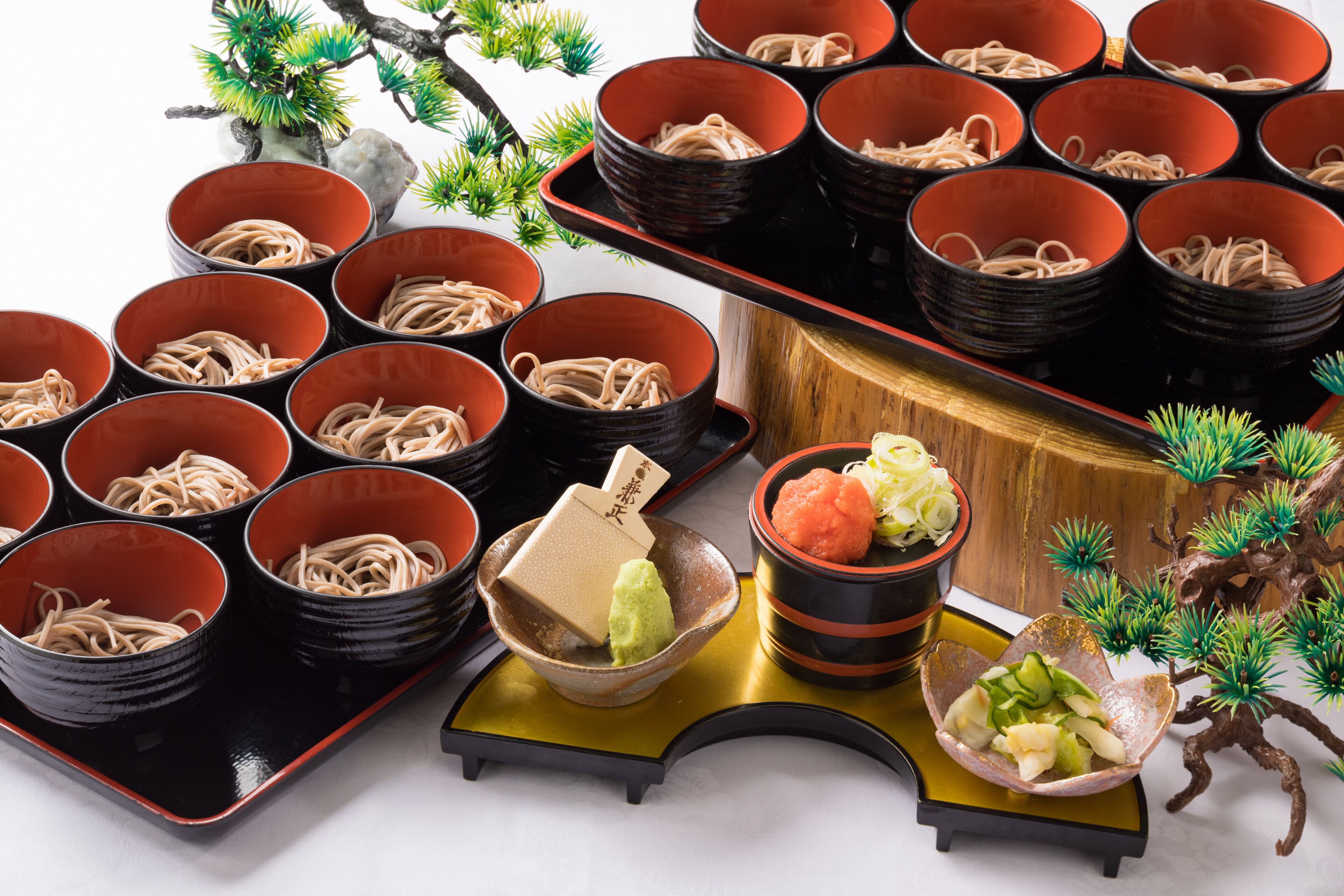 A traditional Japanese meal setup with multiple bowls of noodles, a small dish of wasabi, and a decorative bonsai tree.
