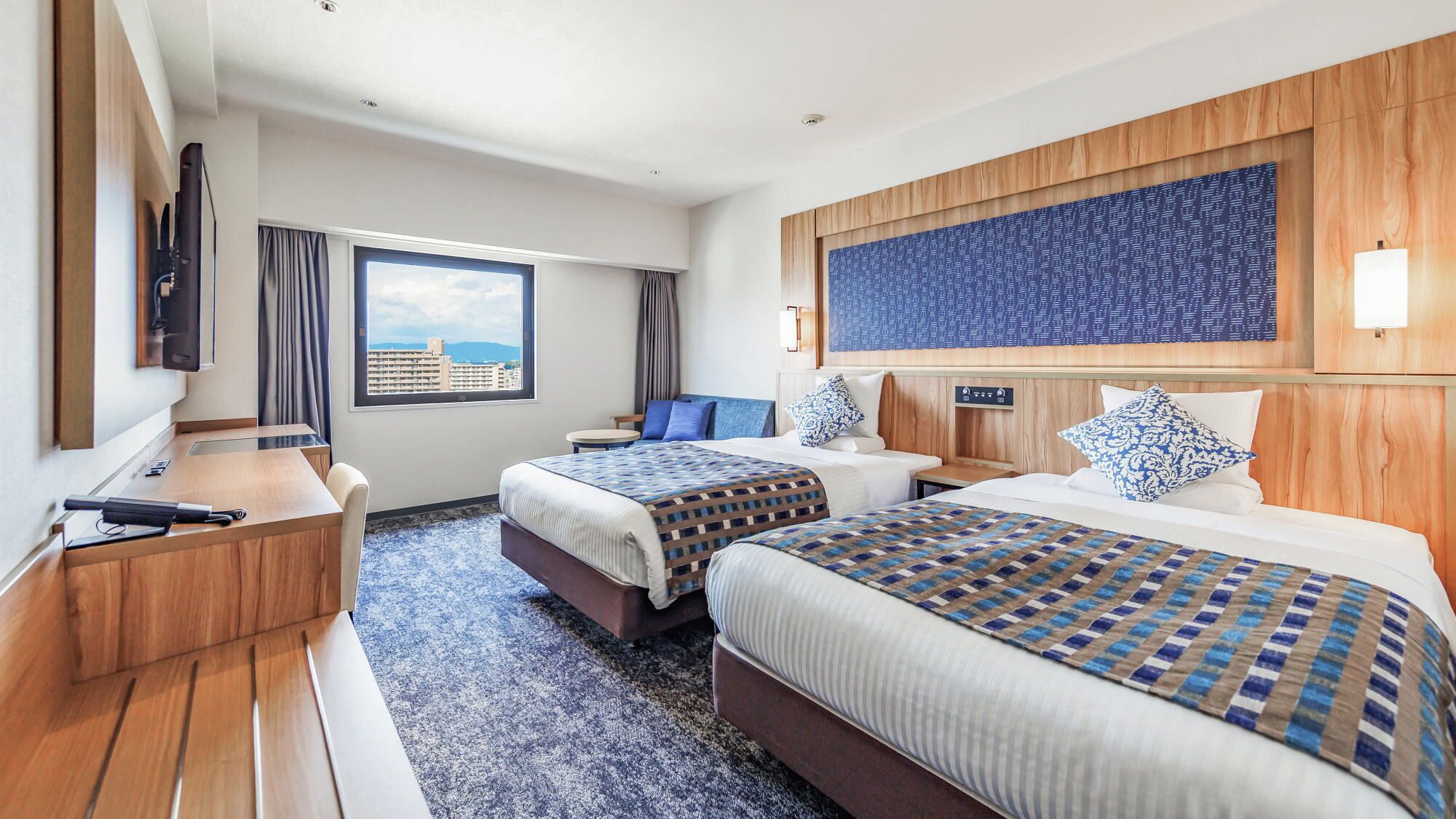 A hotel room with two beds, a desk, a TV, and a large window with a city view.