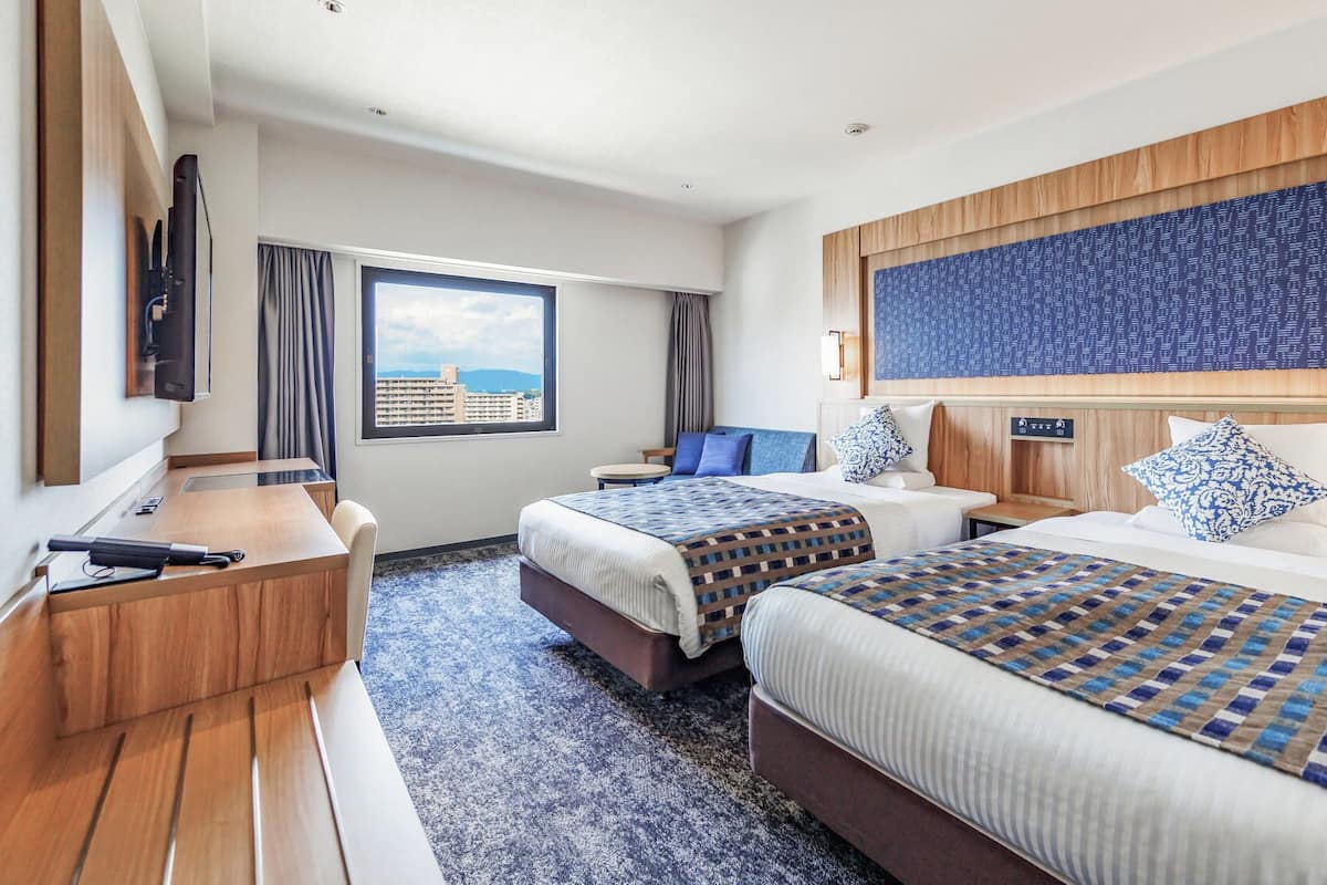 Une chambre d’hôtel avec deux lits, un bureau, une télévision et une grande fenêtre donnant sur la ville.