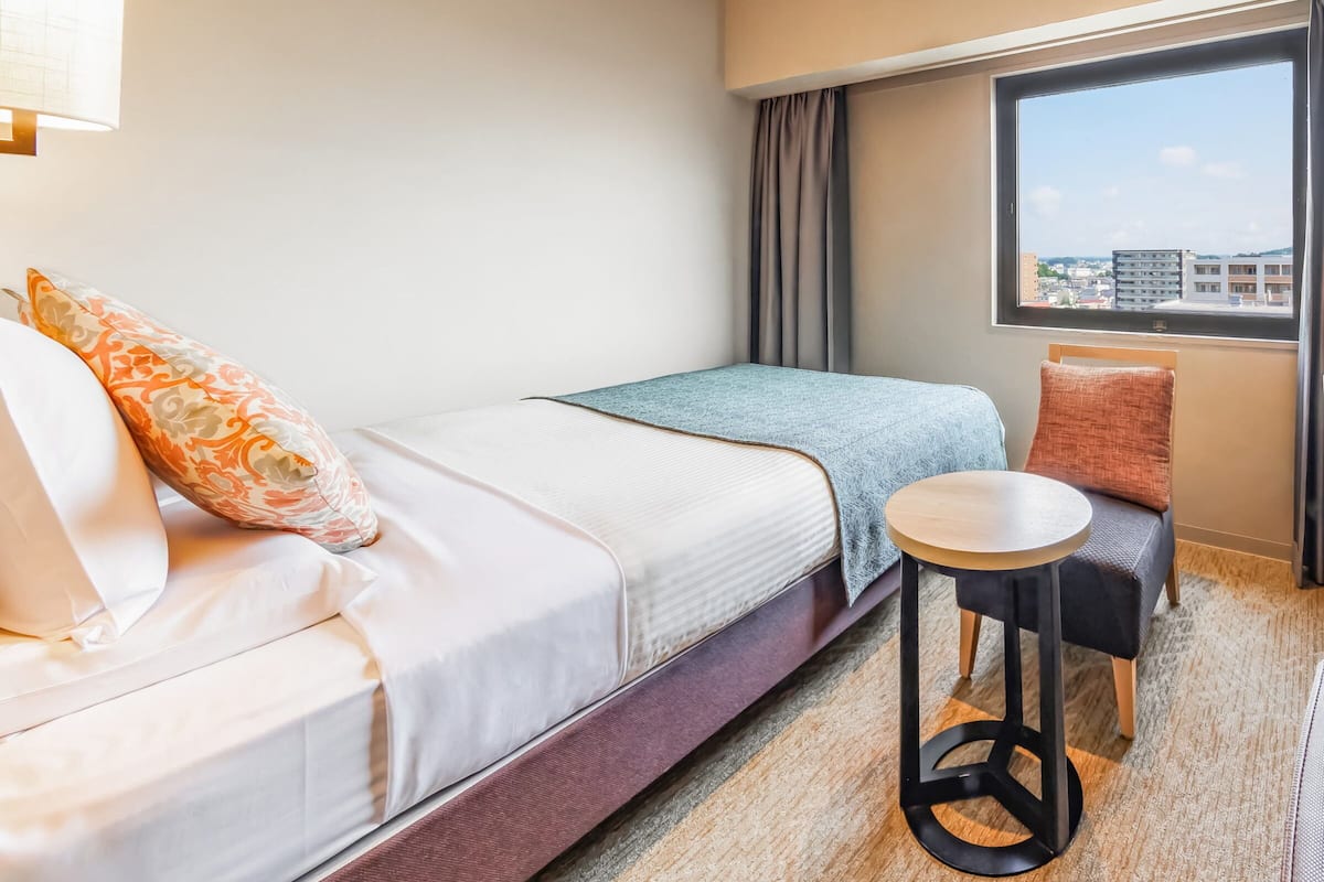 Quarto de hotel com cama, uma mesa redonda pequena, uma cadeira e janela com vista para a cidade.