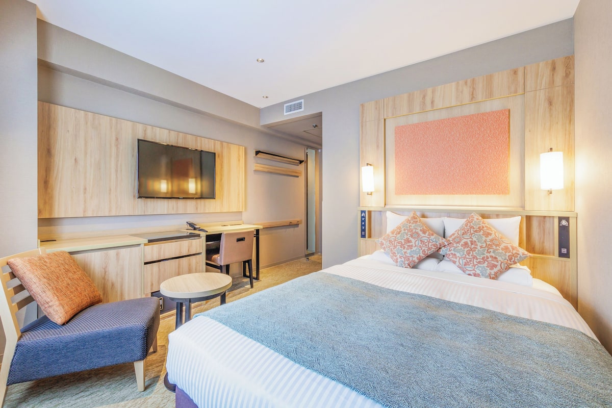 Quarto de hotel com uma cama, uma escrivaninha, uma cadeira e uma televisão.