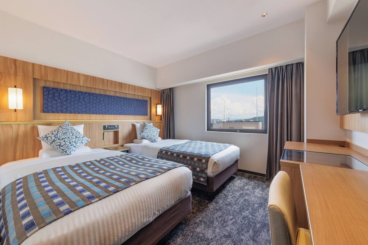 Quarto de hotel com duas camas, uma mesa, uma televisão e uma janela grande com vista.