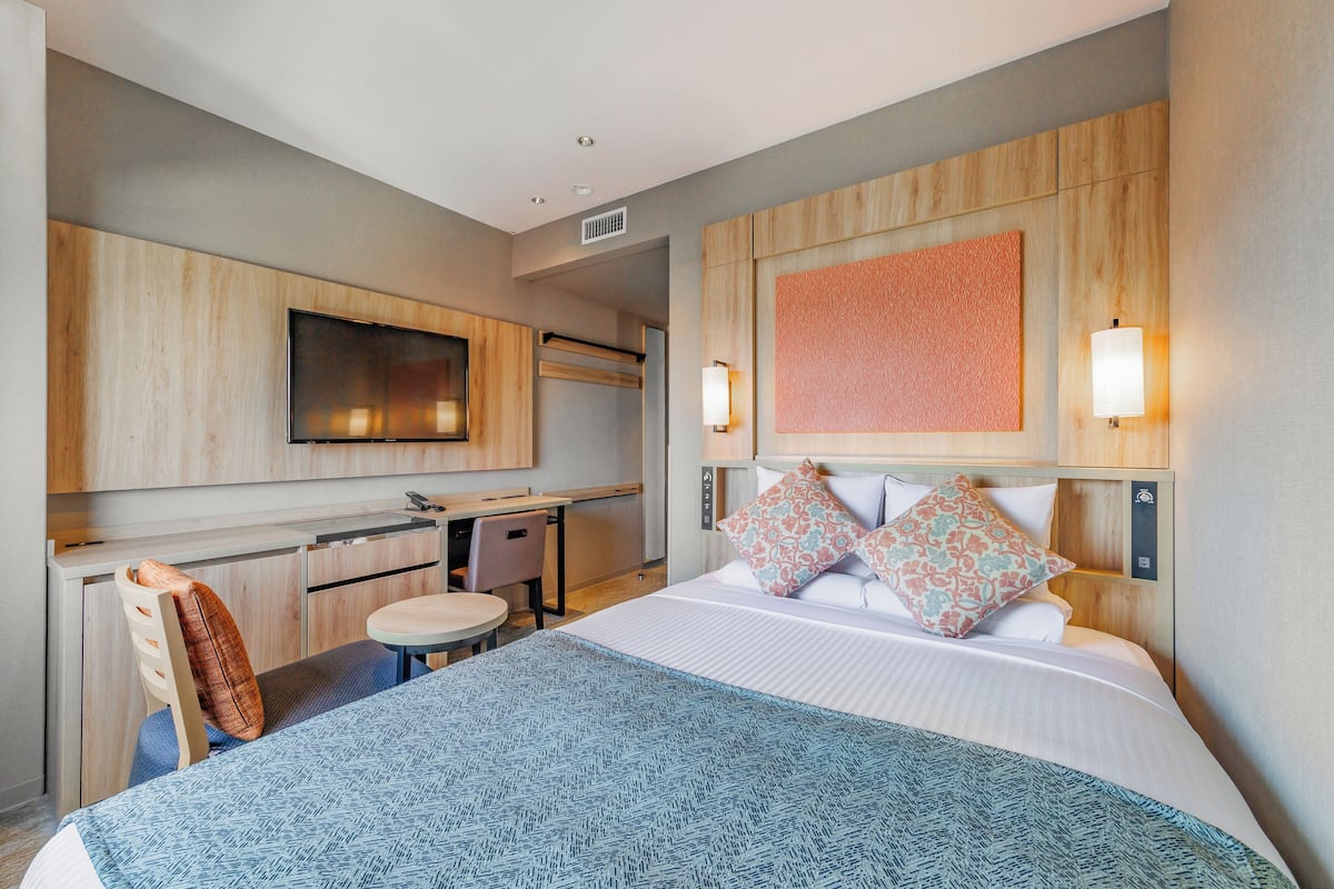 Quarto de hotel com cama, mesa de cabeceira, cadeira, escrivaninha e televisão.