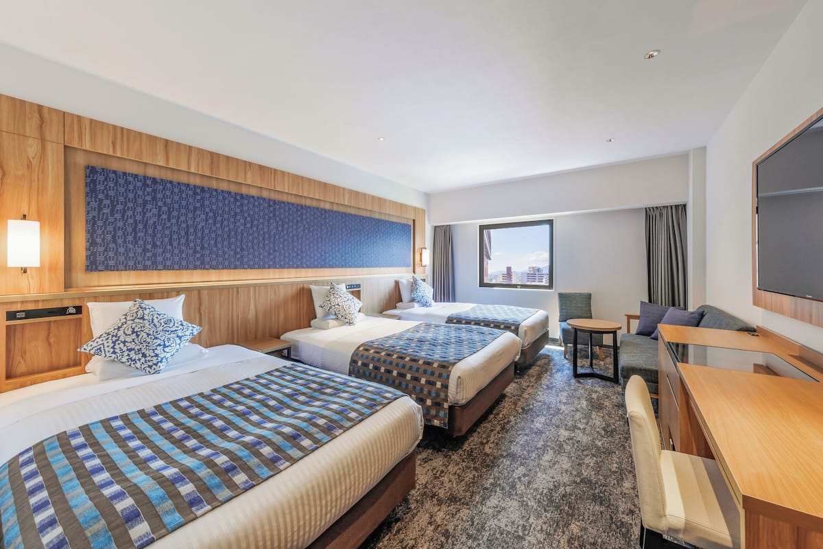 Quarto de hotel com três camas, uma escrivaninha e uma janela grande.