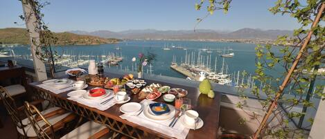 Mesa de jantar posta com pratos, tigelas e talheres, com vista para uma marina e barcos.