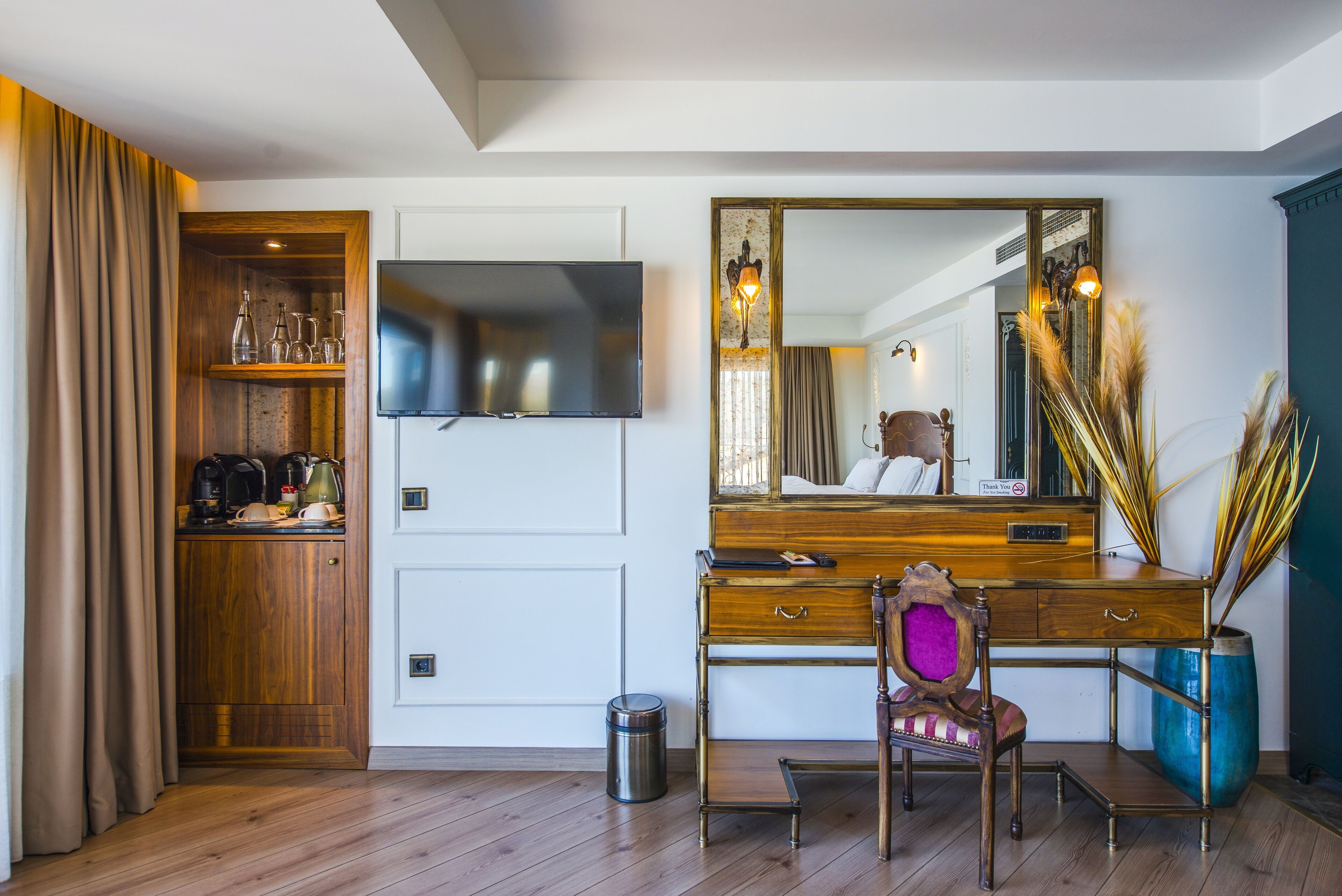 Habitación de hotel con escritorio de madera, una silla, televisor, espejo, mueble pequeño y un cubo de basura.