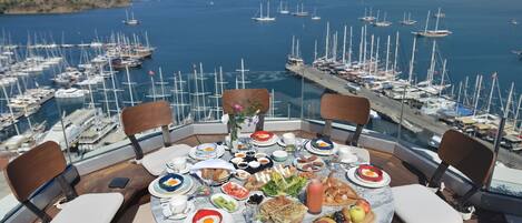A round table with a variety of food and drinks, surrounded by chairs, overlooking a marina with boats.
