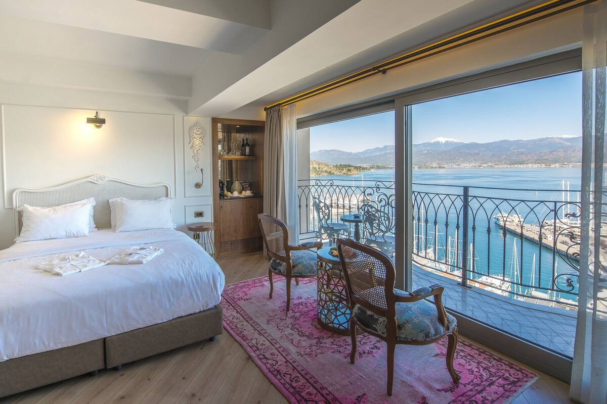 A bedroom with a large bed, a wooden side table, a small table with chairs, and a view of the sea and mountains through a balcony.