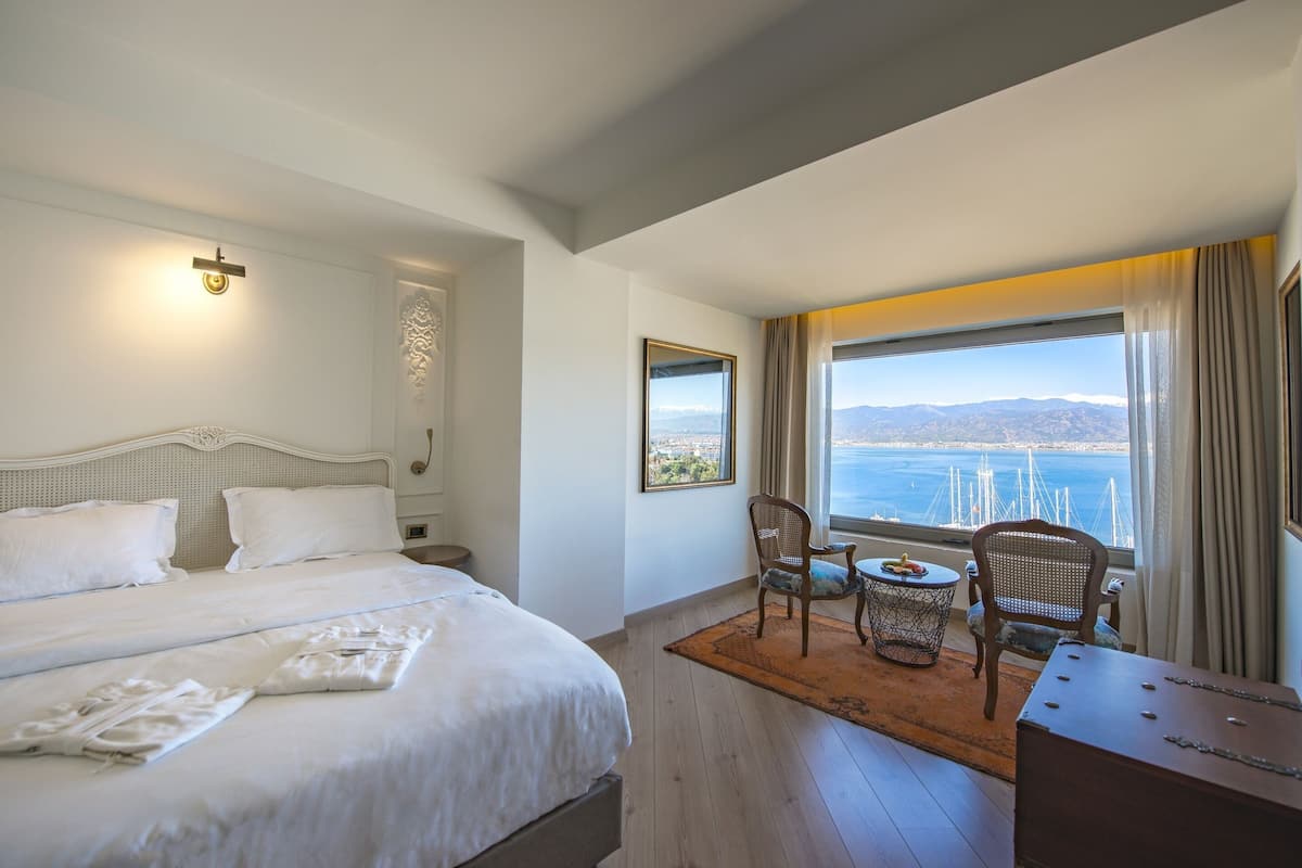 A hotel room with a large bed, a view of the sea, and a dining area with a table and chairs.