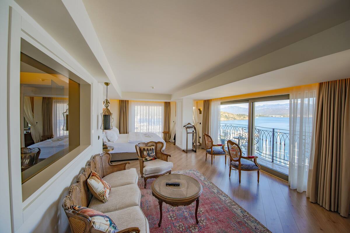 A spacious hotel room with a large bed, a sitting area with a sofa and chairs, a small table, and a view of the sea through large windows.