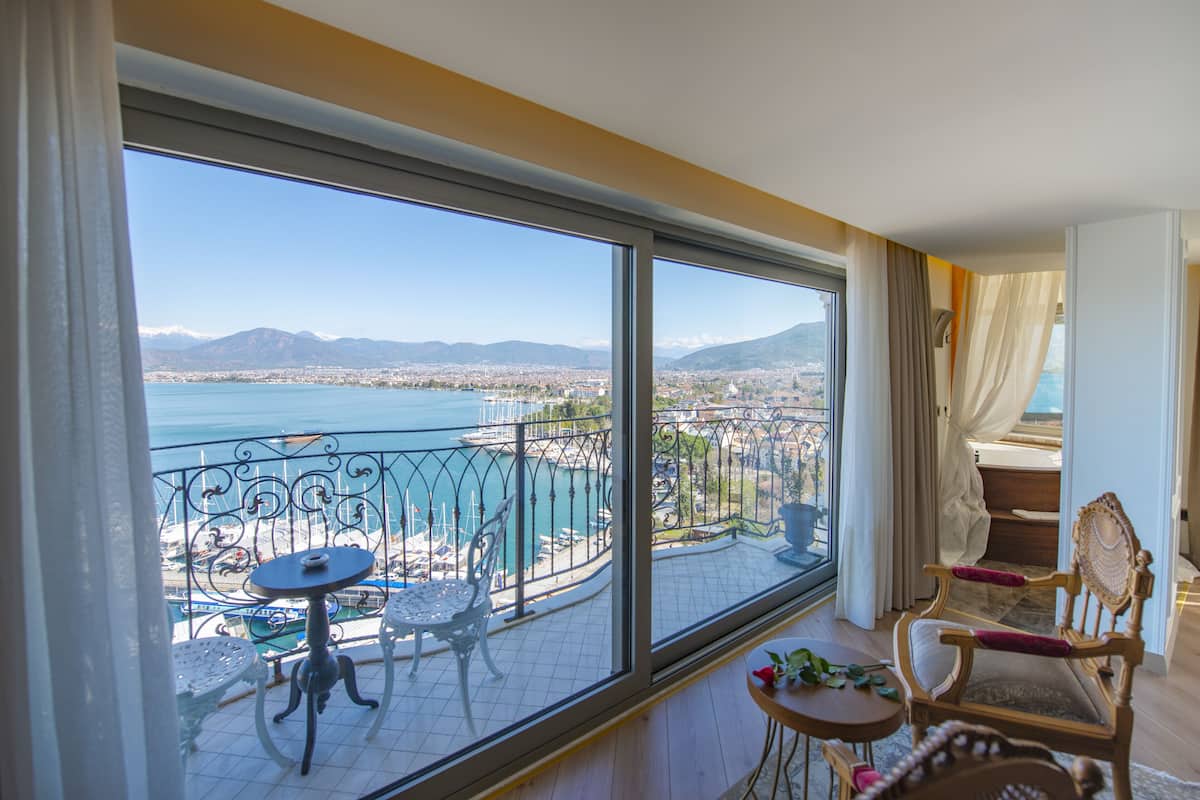 A balcony with a table and chairs, a view of a body of water and mountains, and a cityscape in the distance.
