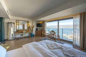 A hotel room with a large bed, a desk, a chair, a TV, and a view of the sea.