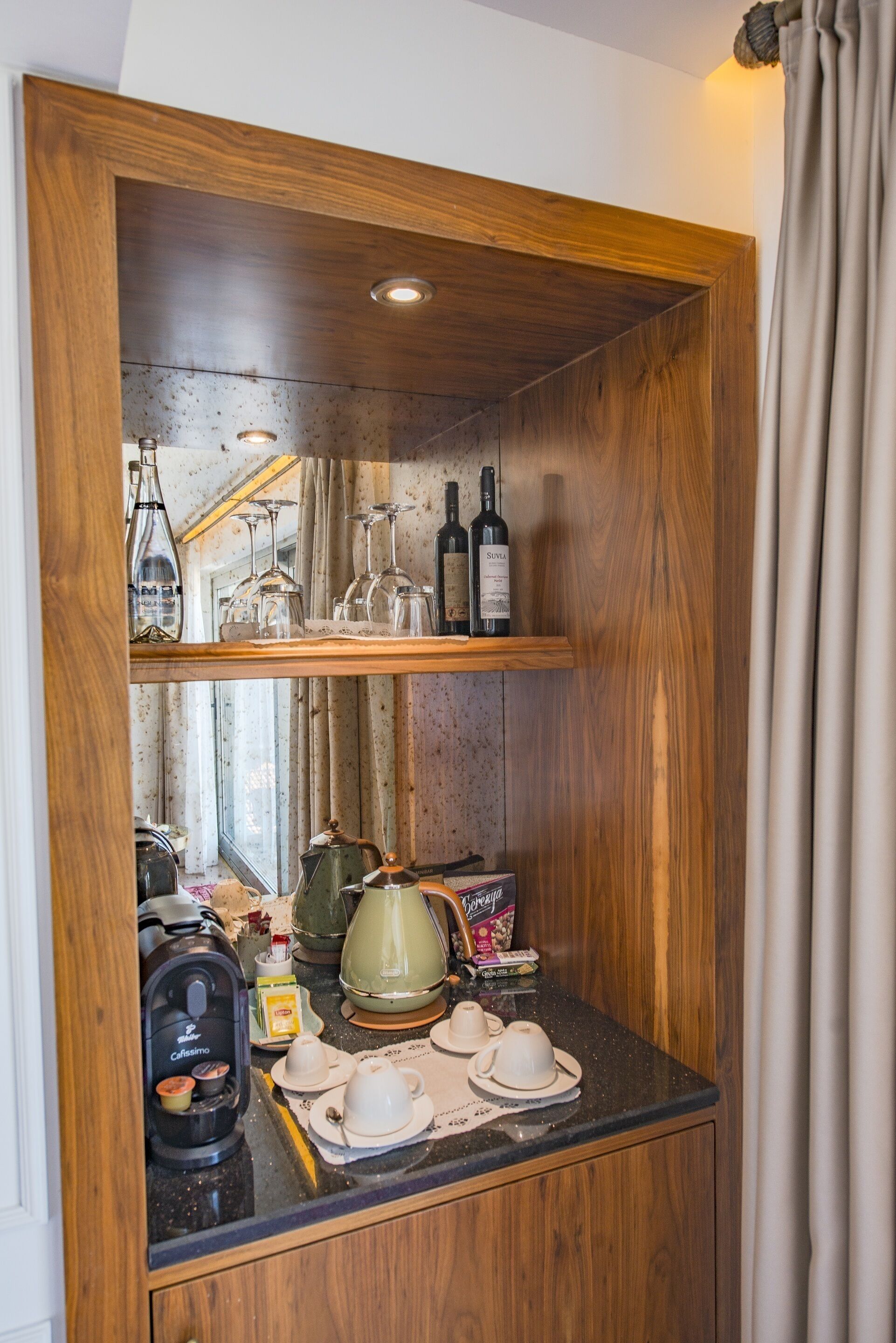 A wooden cabinet with shelves holding wine bottles and glasses, a coffee maker, and a teapot.