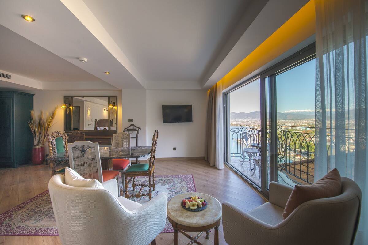 A spacious living room with a large window offering a city view, a dining area with a table and chairs, and a cozy seating area with a small table.