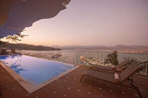 Ein Infinity-Pool mit Liegestühlen, Blick auf einen Yachthafen und ferne Berge.