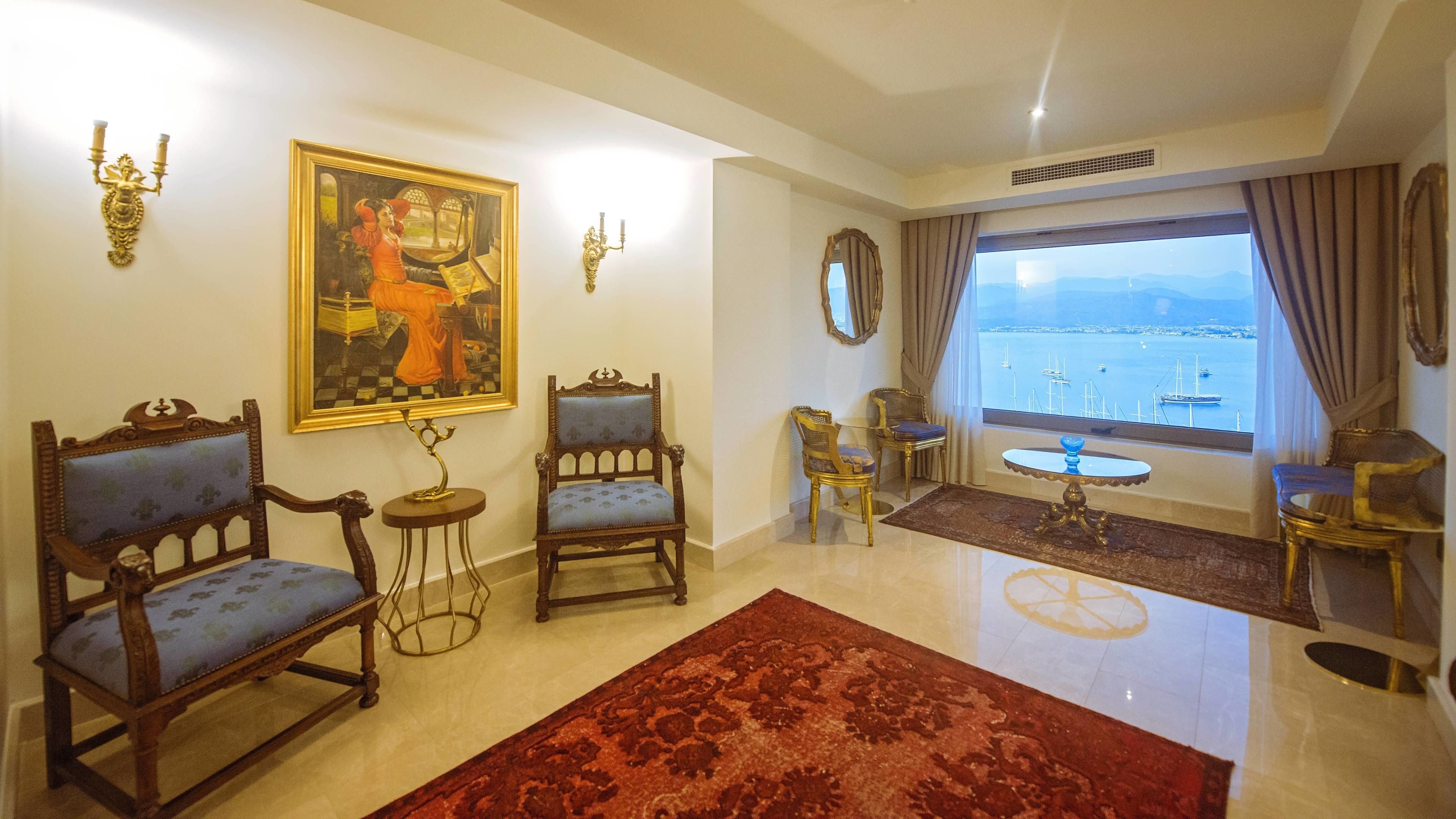 A spacious living room with a large window offering a view of the sea. There are two ornate chairs, a small table, and a red rug on the floor.