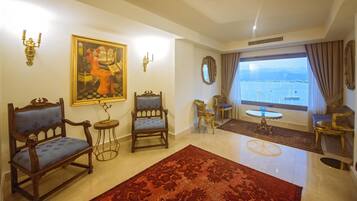 A spacious living room with a large window offering a view of the sea. There are two ornate chairs, a small table, and a red rug on the floor.