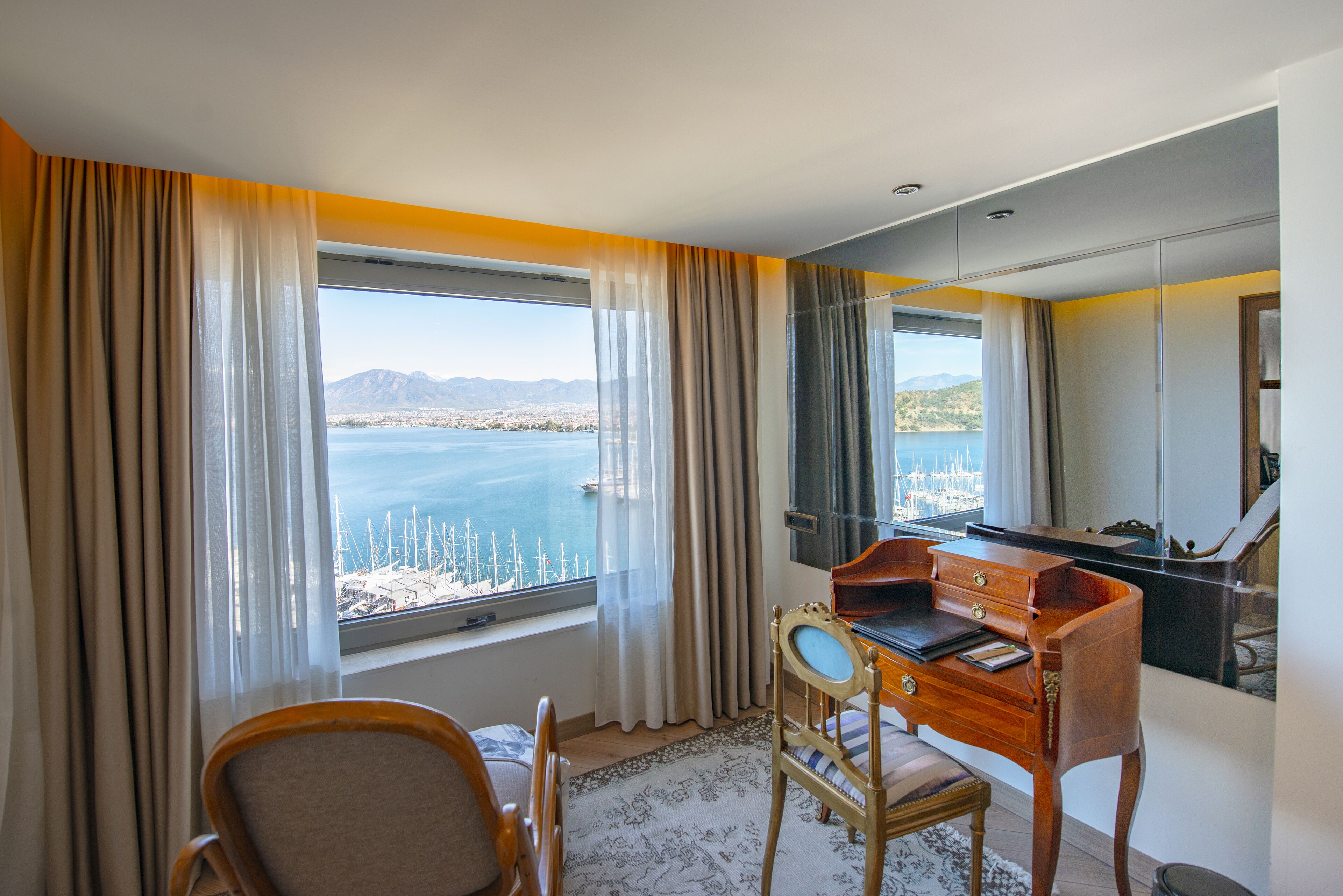 A hotel room with a desk, chair, and a large window offering a view of a body of water and distant mountains.