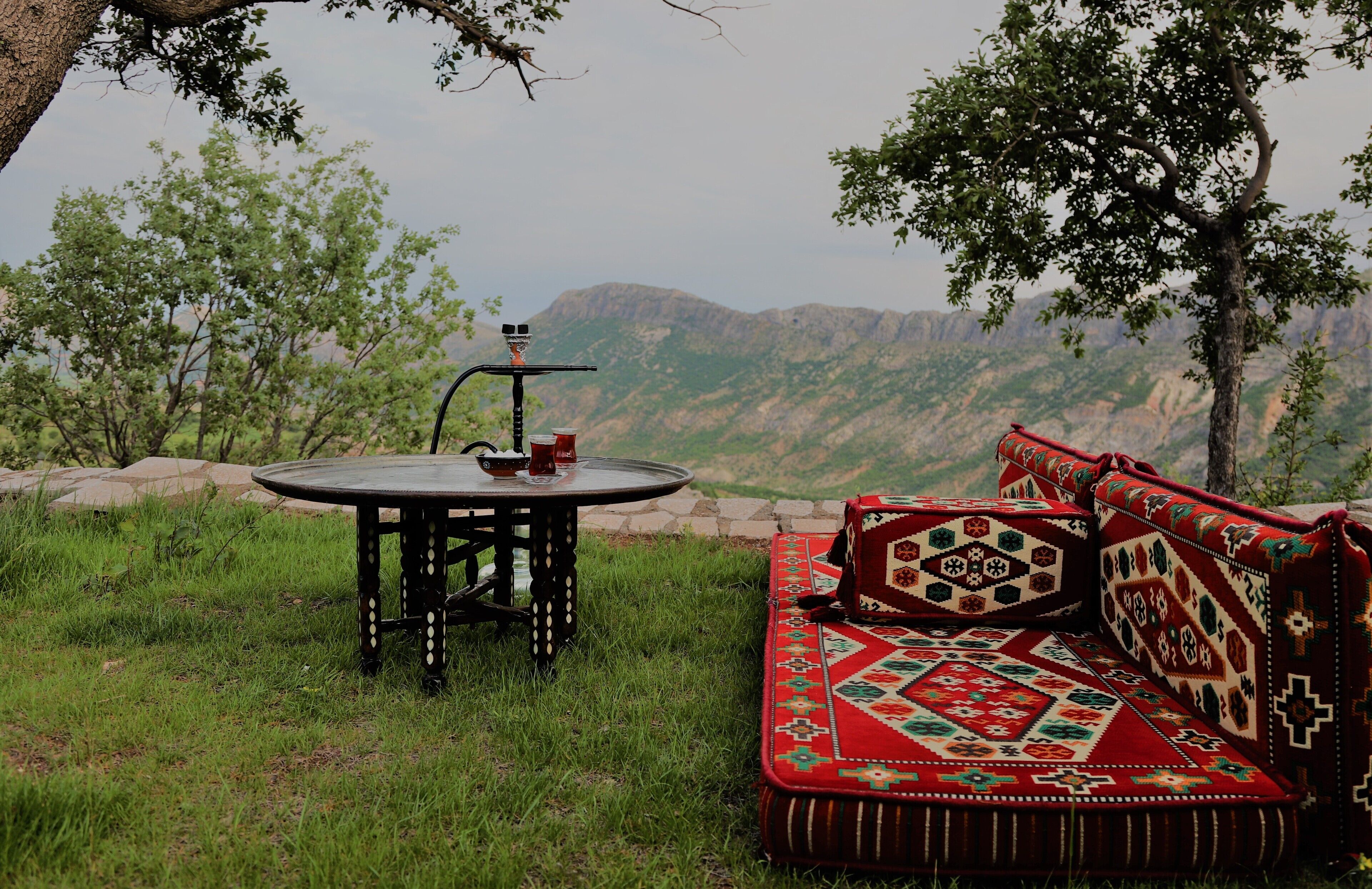 Una mesa redonda con un narguile y dos tazas, un sofá rojo con estampado, y un paisaje montañoso al fondo.