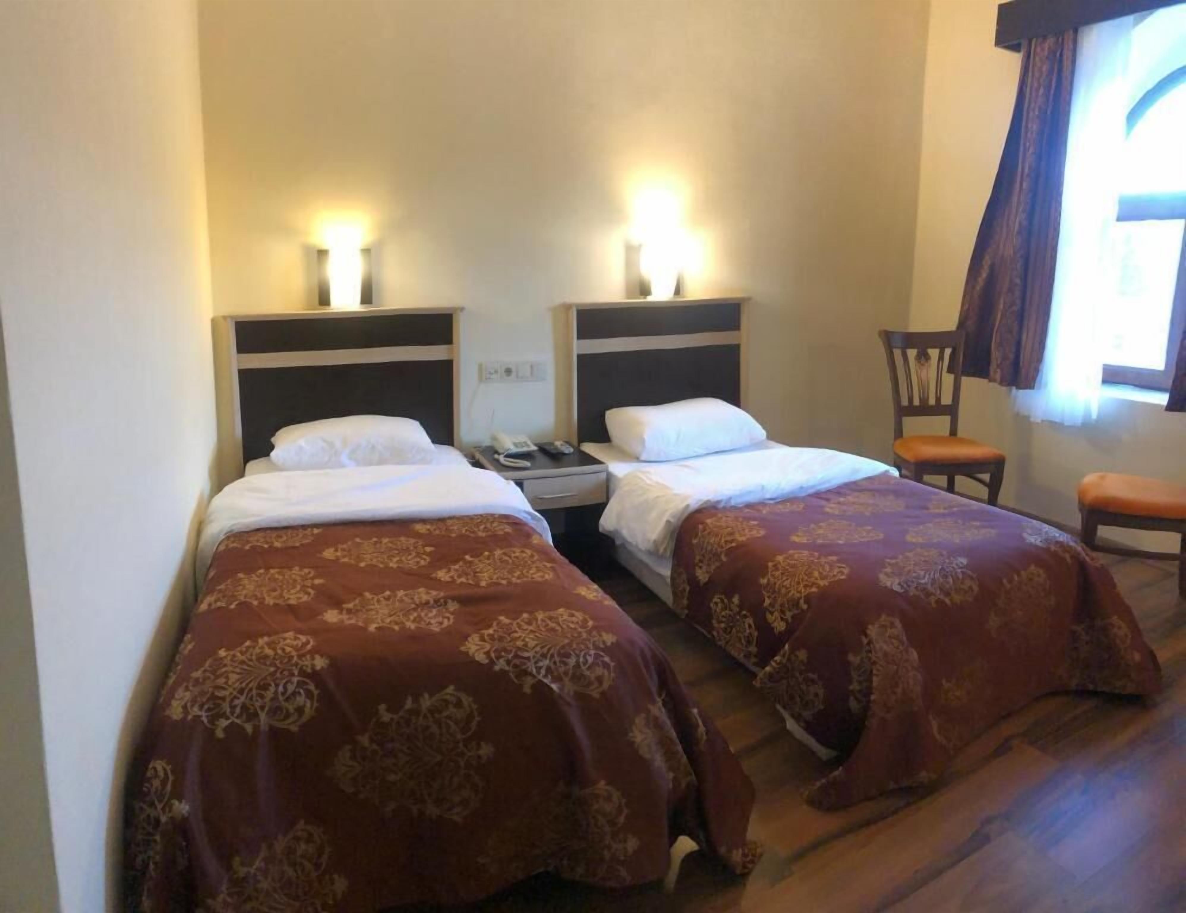 A hotel room with two beds, each with a headboard and bedside lamps. There is a chair and a window with curtains.