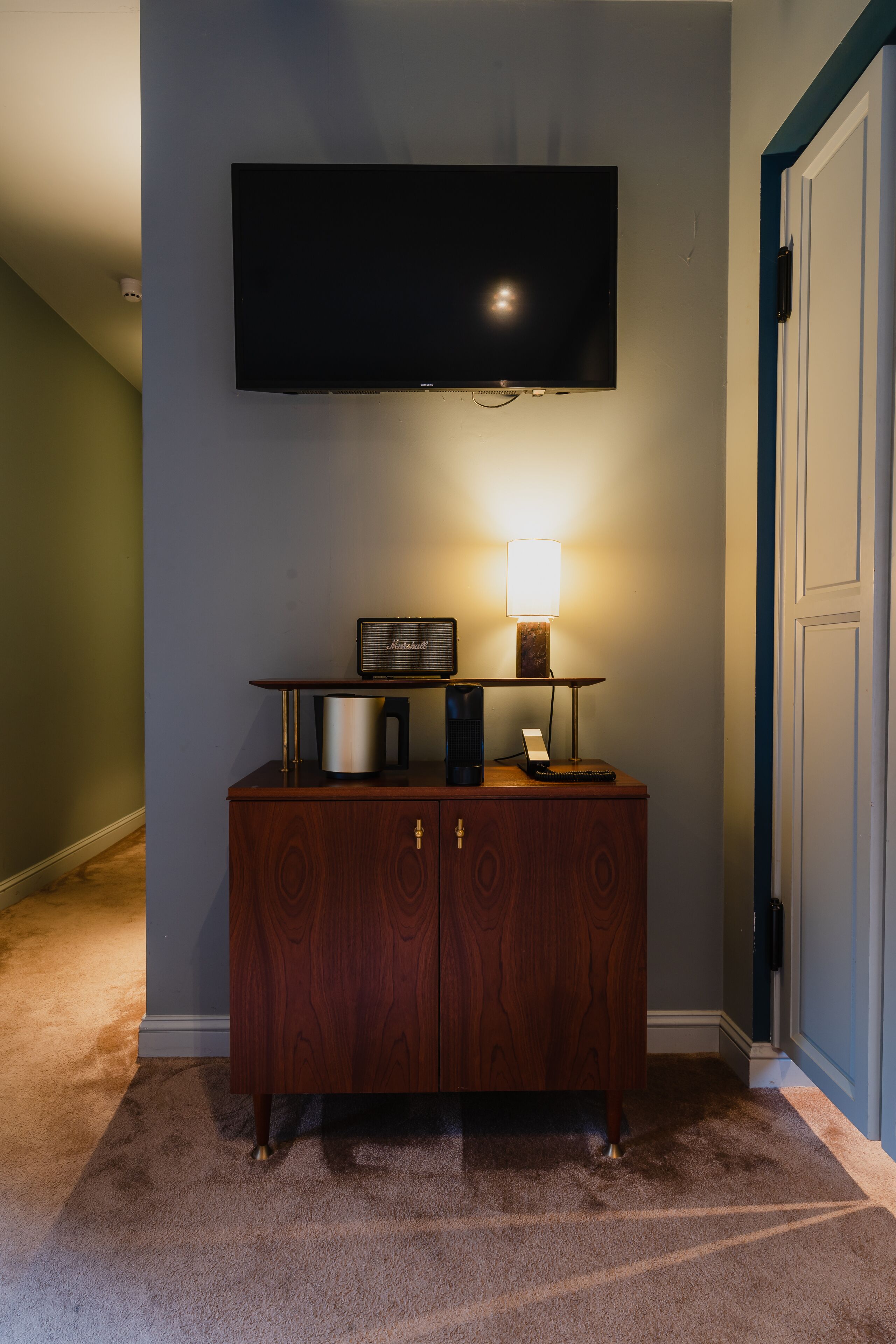 Un mueble de madera con un televisor de pantalla plana montado encima, una lámpara y una mesita con un teléfono y una cafetera.