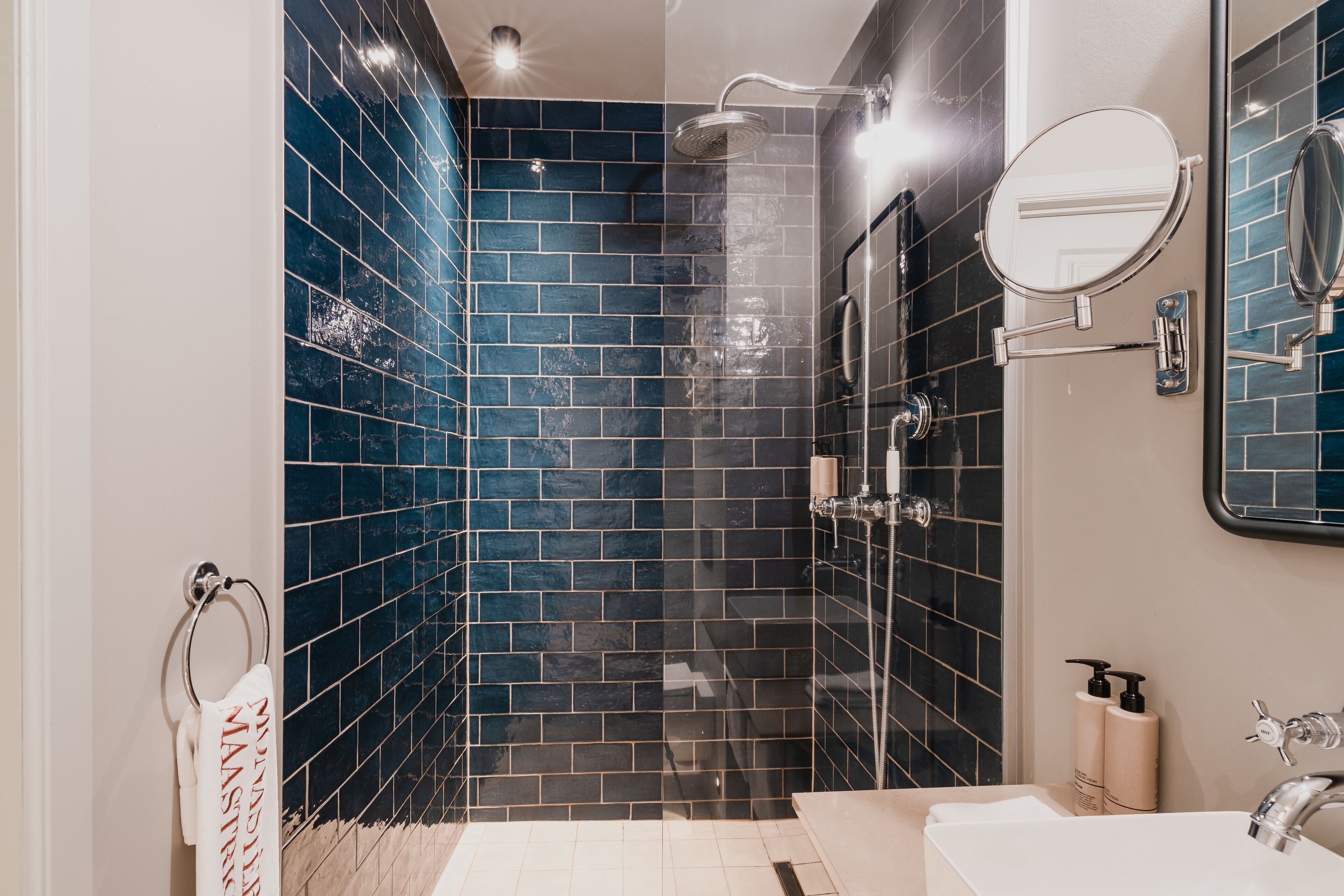 A bathroom with dark blue tiled walls, a glass-enclosed shower, a white sink, and a mirror.