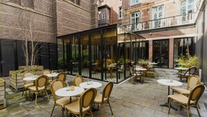Un espace extérieur aménagé en terrasse, avec des chaises et des tables en osier, entouré de bâtiments en briques.