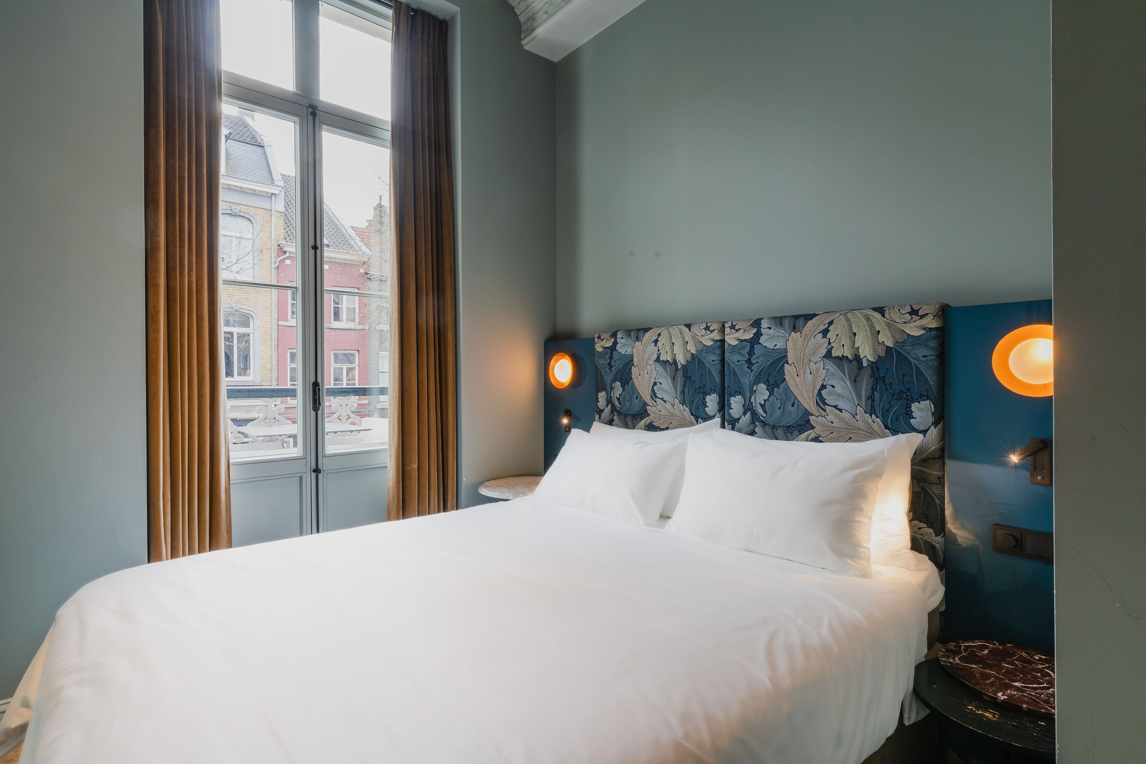 Una habitación de hotel con una cama grande, una ventana con cortinas y una lámpara fijada a la pared.