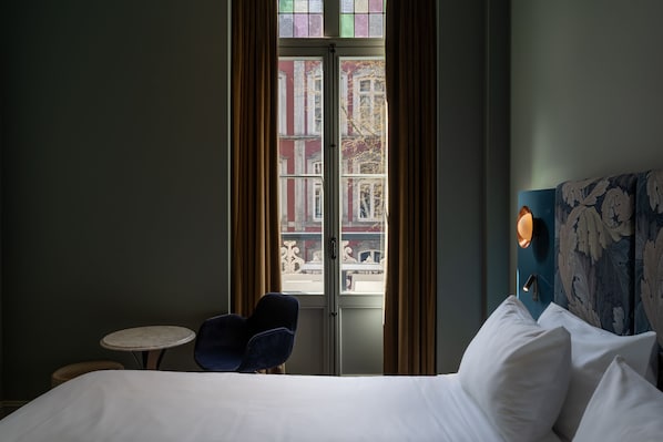 Habitación de hotel con cama, una mesita redonda, una silla y vistas a edificios a través de una ventana con cortinas.