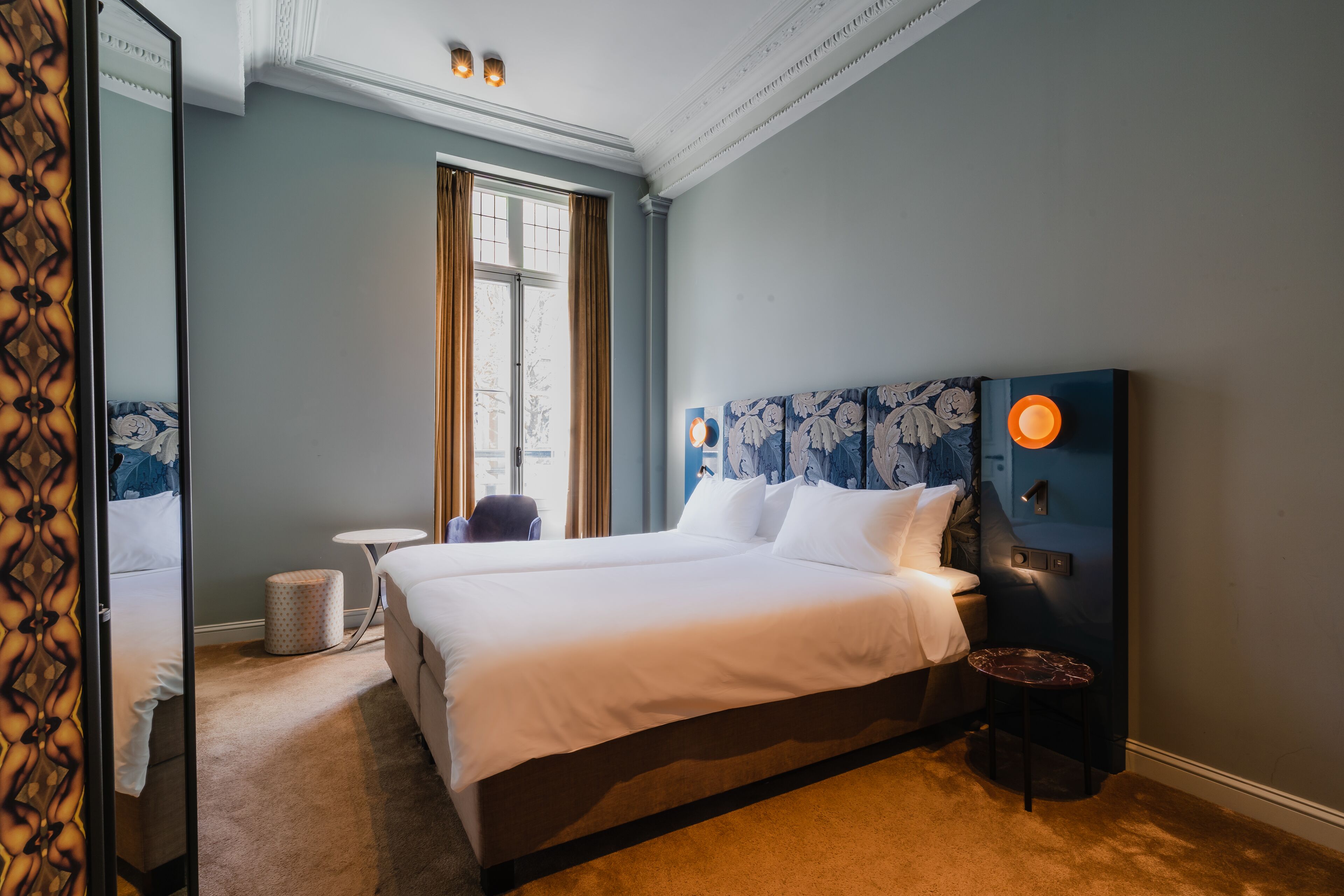 Ein Hotelzimmer mit einem großen Bett, einem kleinen runden Tisch, einem Sessel, einem Mülleimer und einem Fenster mit Vorhängen.