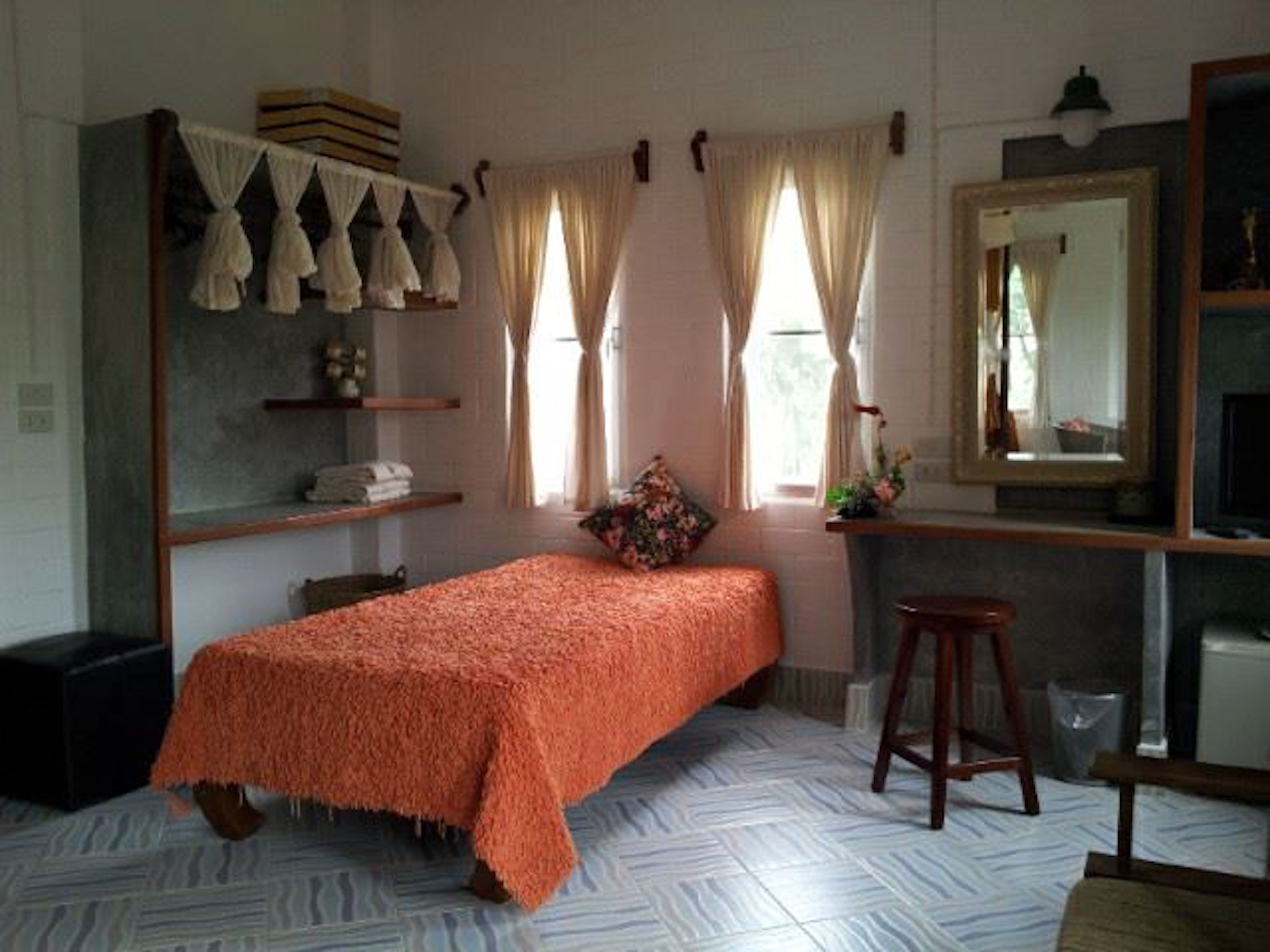 Quarto com uma cama, um banquinho, um espelho e uma janela com cortinas.