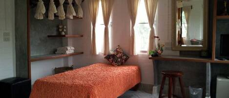 Quarto com uma cama, um banquinho, um espelho e uma janela com cortinas.