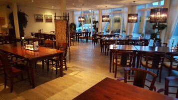 Um restaurante amplo com mesas e cadeiras de madeira, janelas grandes e luminárias pendentes.