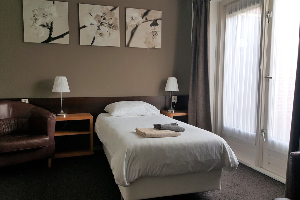 A hotel room with a bed, two bedside lamps, a brown leather chair, a wooden nightstand, and three framed pictures on the wall.