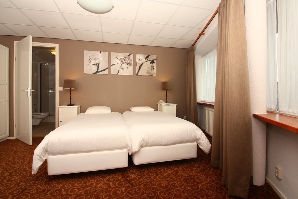 A double bed with a white comforter, two bedside tables with lamps, a window with curtains, and a door leading to a bathroom.