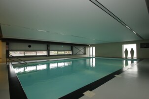 Piscina interna com água cristalina, piso azulejado e janelas que permitem a entrada de luz natural.