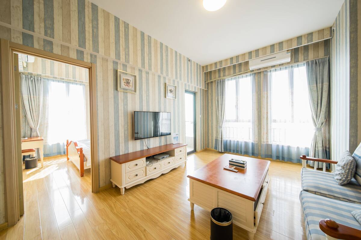 Un salon spacieux doté d’un plancher en bois, d’un canapé, d’une table basse, d’un téléviseur et d’une fenêtre avec des rideaux.