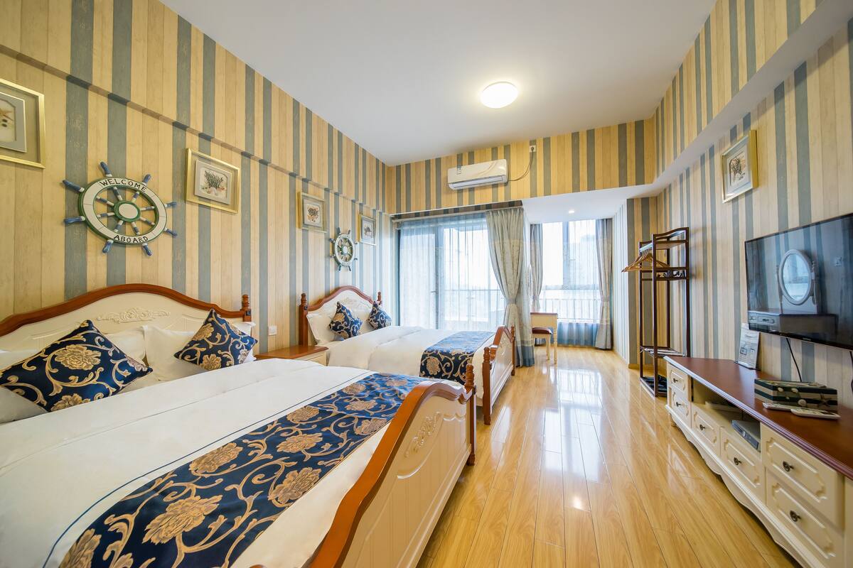 Une chambre d’hôtel avec un papier peint à rayures, deux lits dotés de têtes de lit décorées, une commode en bois, un téléviseur et une fenêtre avec des rideaux.