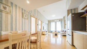 A spacious living area with a wooden dining table and chairs, a kitchen with a refrigerator, and a sofa in the background.