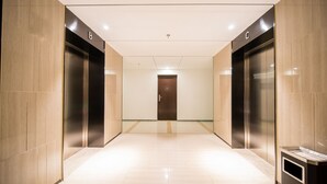 A modern elevator lobby with two elevators marked B and C, a wooden door at the end, and a trash bin to the right.