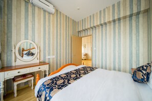 Een slaapkamer met gestreepte behang, een houten dressoir met spiegel, een bed met een bedspread met patroon, en een deur die naar een andere kamer leidt.
