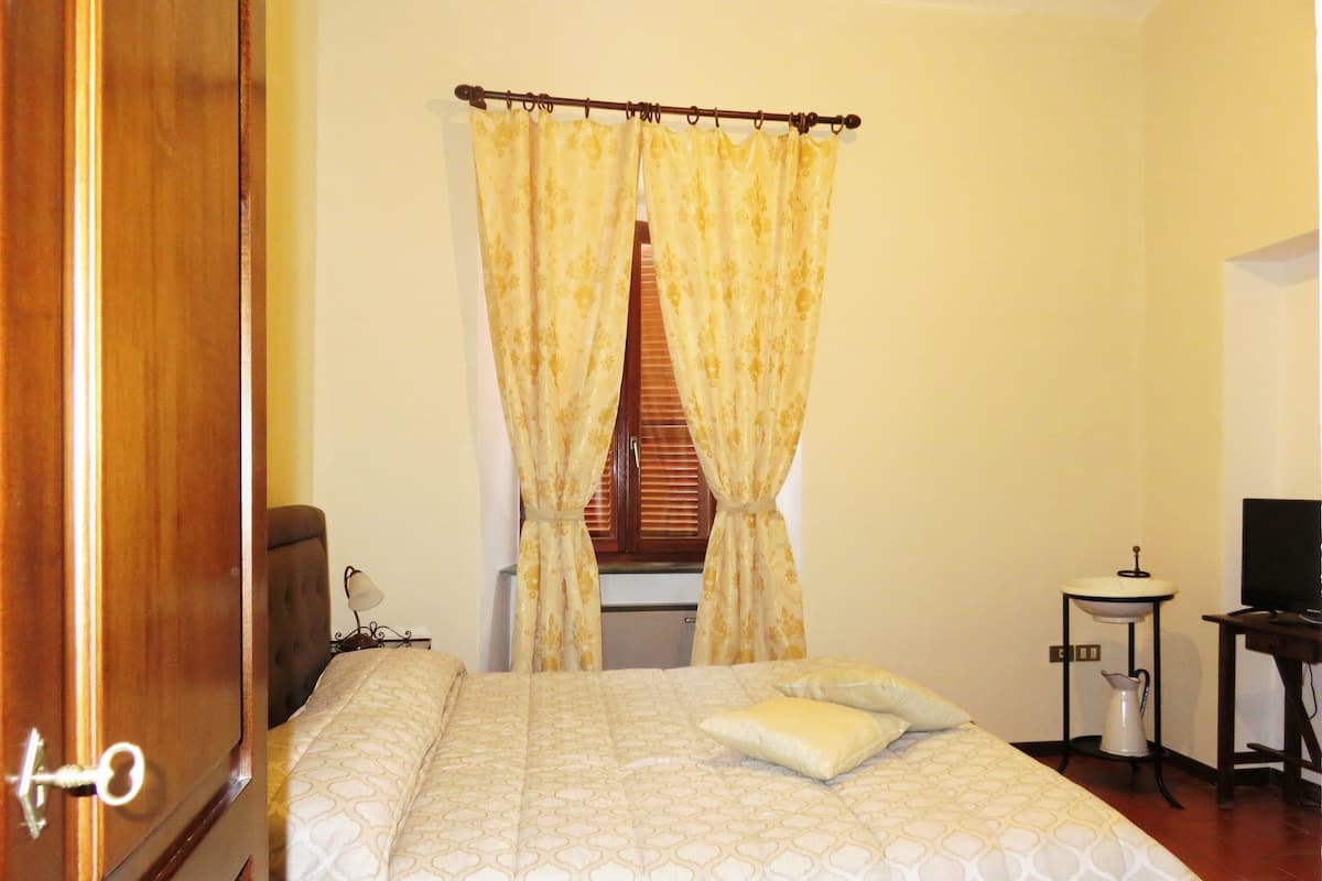 Un dormitorio con cama, cortinas, una ventana con persianas, una mesita de noche con lámpara y un televisor.