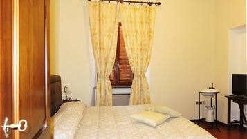 A bedroom with a bed, curtains, a window with blinds, a bedside table with a lamp, and a television.