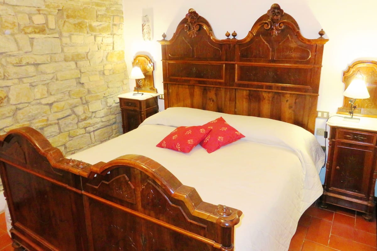 Una cama de madera con cabecera, dos mesitas de noche con lámparas, una pared de piedra y suelo de baldosas.